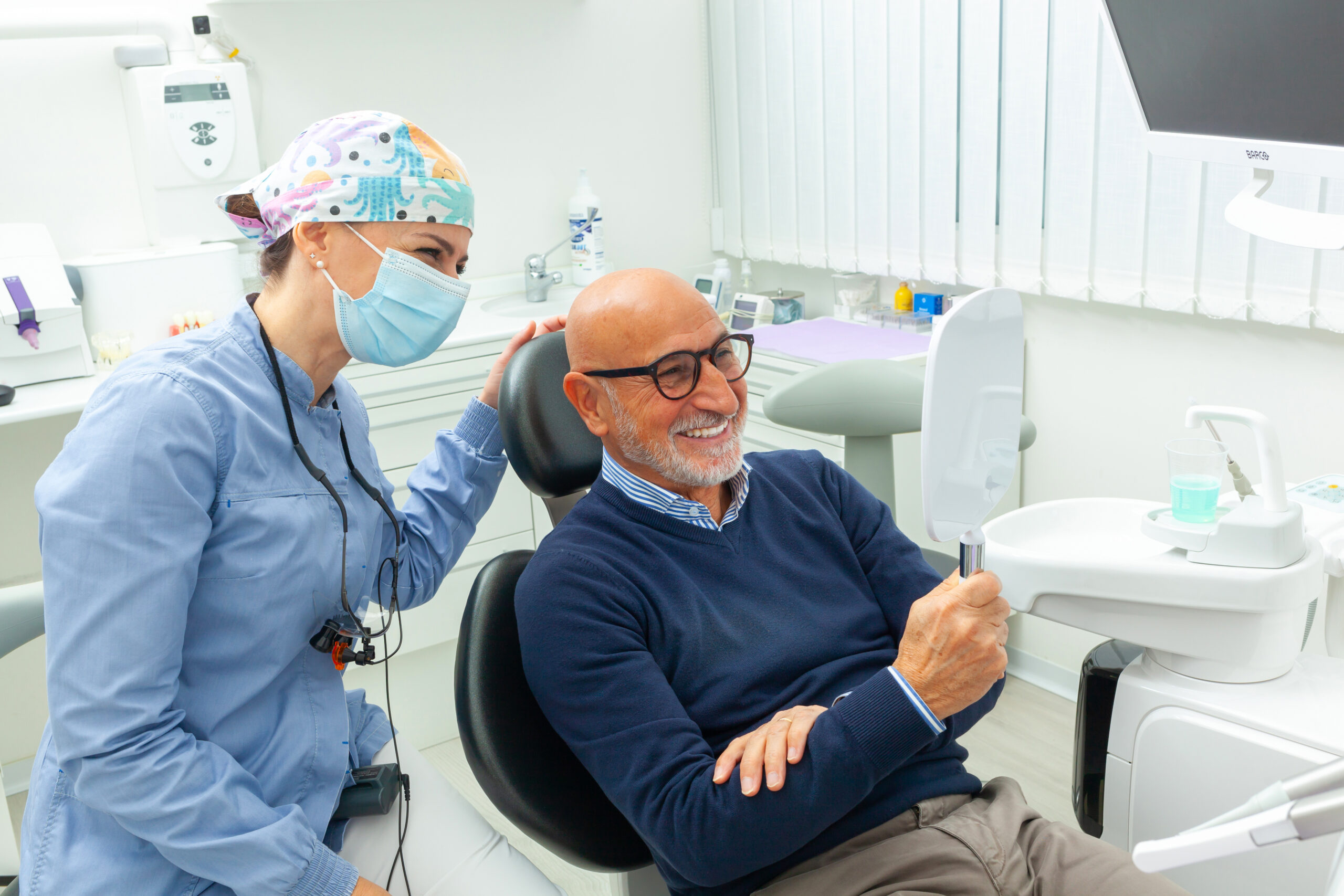 Smiling senior man looking at his new smile in the mirror at the dental clinic, assisted by a kind female dentist
