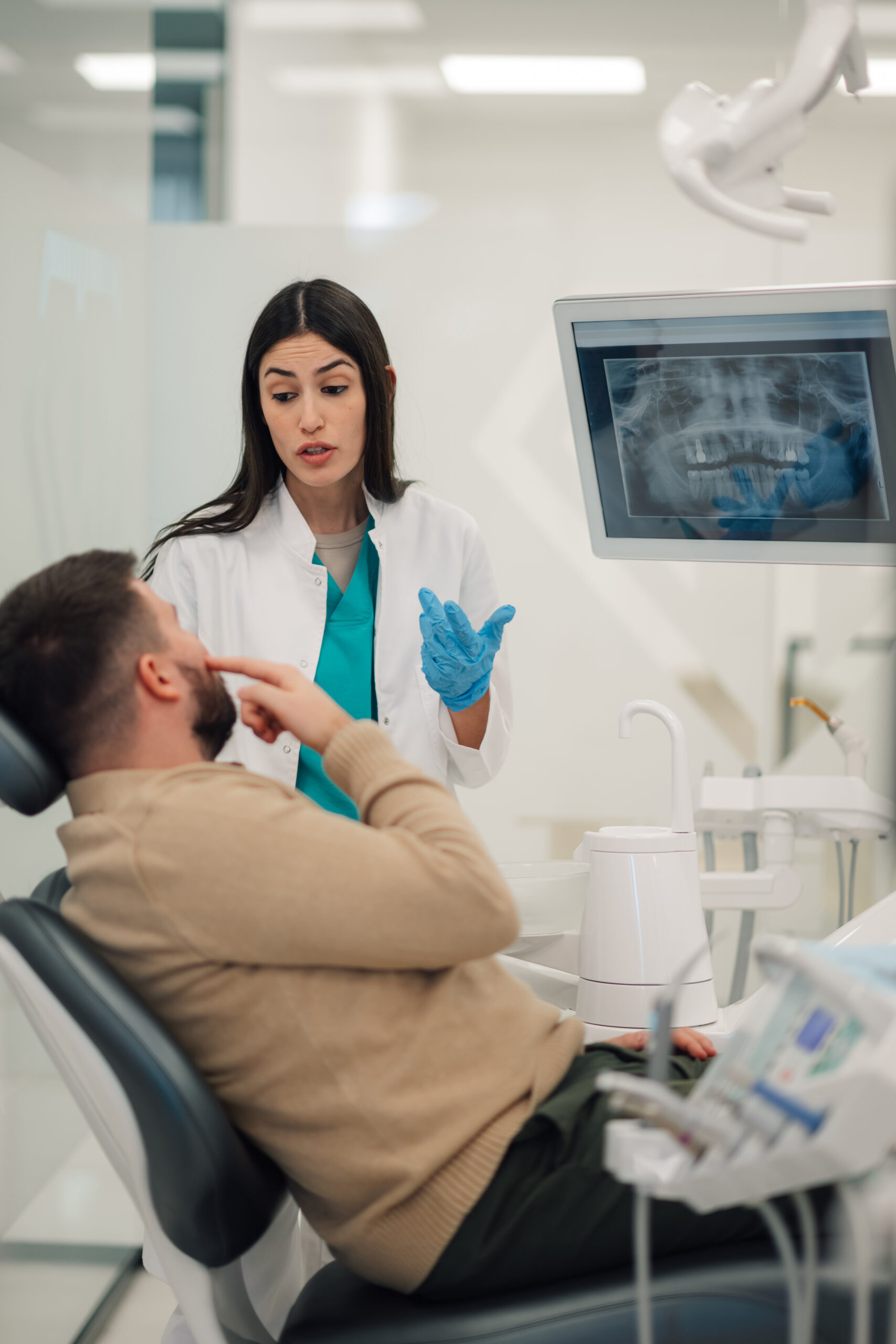Female dentist wearing blue gloves is explaining a dental x ray displayed on a monitor to a male patient sitting in a dental chair, discussing his dental health and treatment options