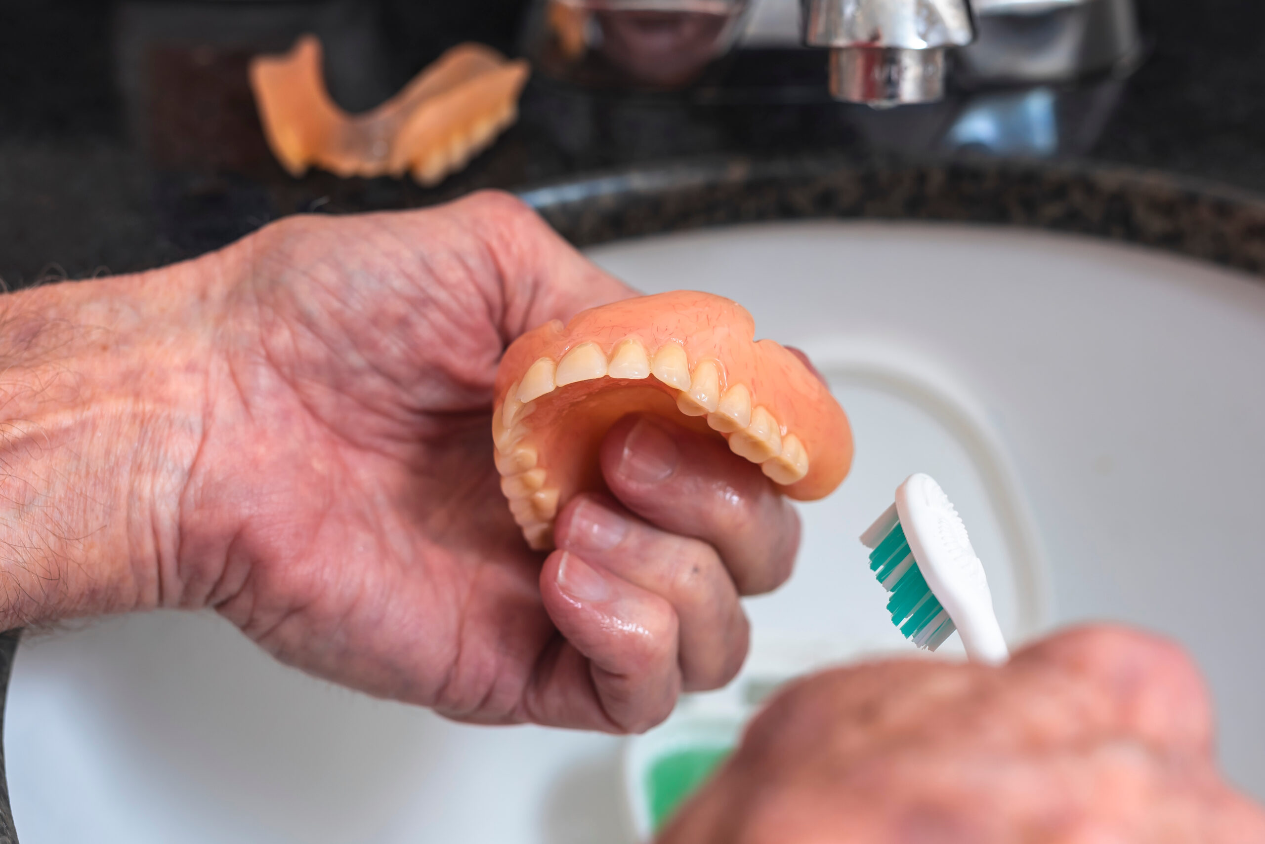 An elderly person holds a full upper denture while cleaning it with a toothbrush over a denture soaking case filled with green cleaning solution, illustrating proper hygiene care.