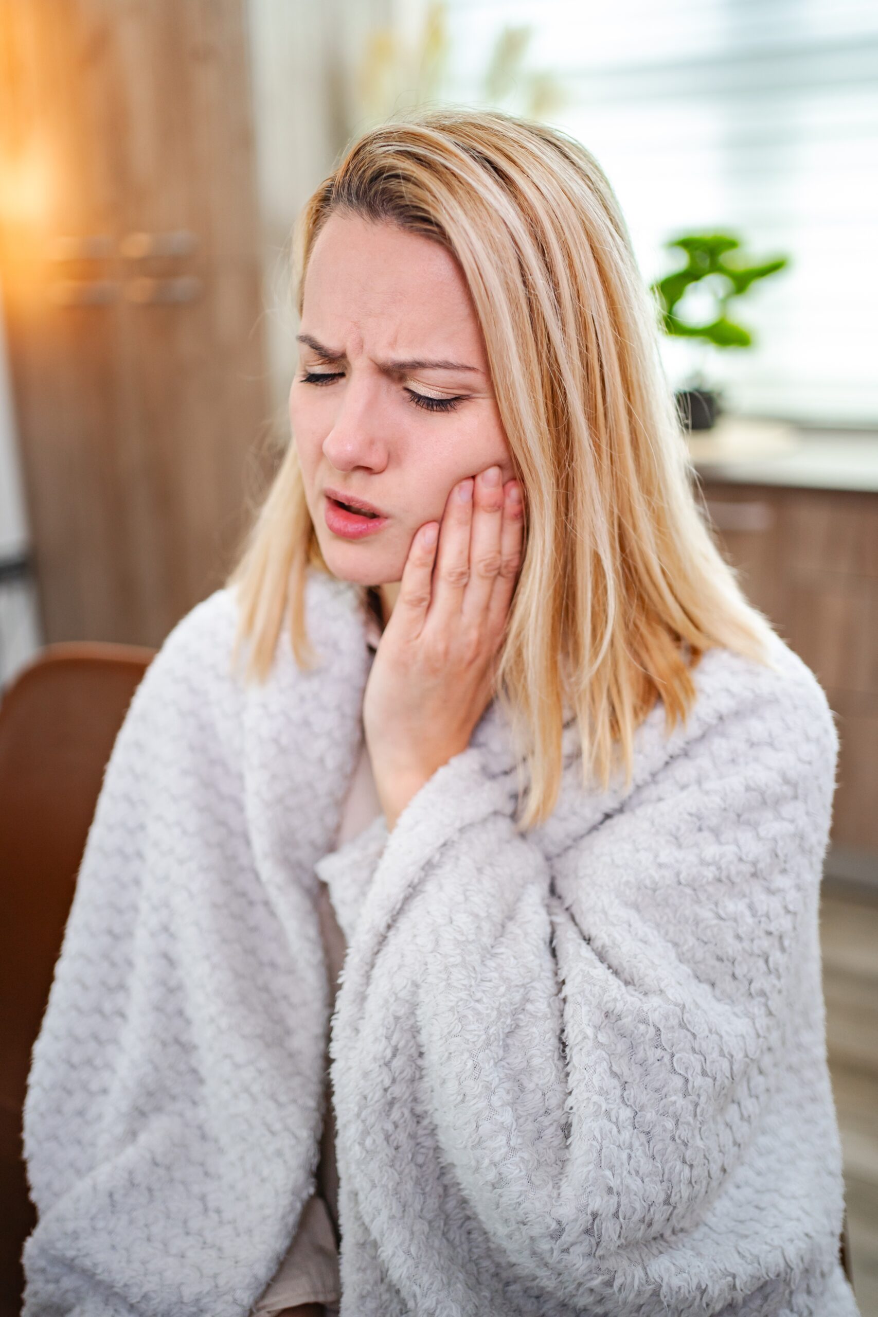 A young woman clutches her jaw, showing discomfort while sitting indoors.