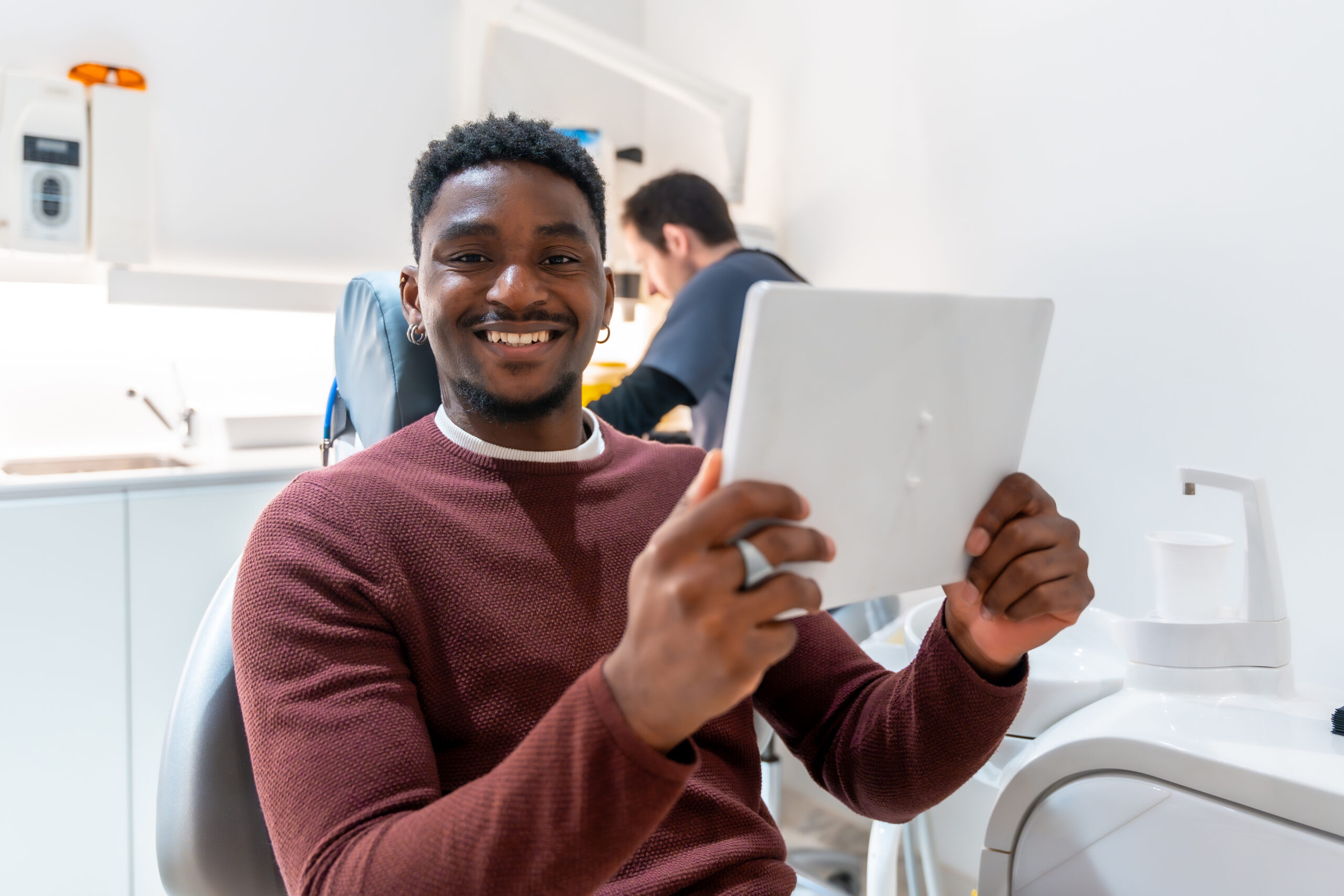Smiling patient using digital tablet, possibly reviewing treatment plan or x rays, while dentist works in background of modern dental clinic