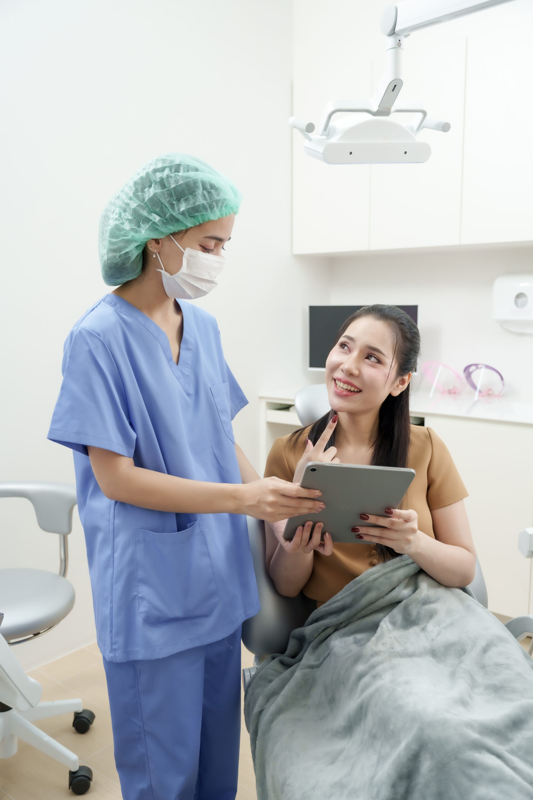 asian female dental assistant in blue uniform consults with asian adult female patient who points to tooth while discussing dental issue using tablet inside clean white dental clinic