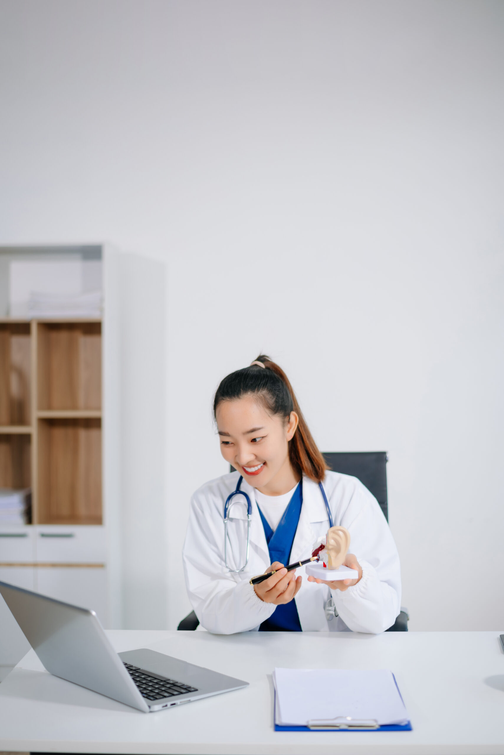 Smiling young Asian dentist or doctor demonstrates a tooth model at her office desk with a laptop showcasing real people, dental education, and professional care.