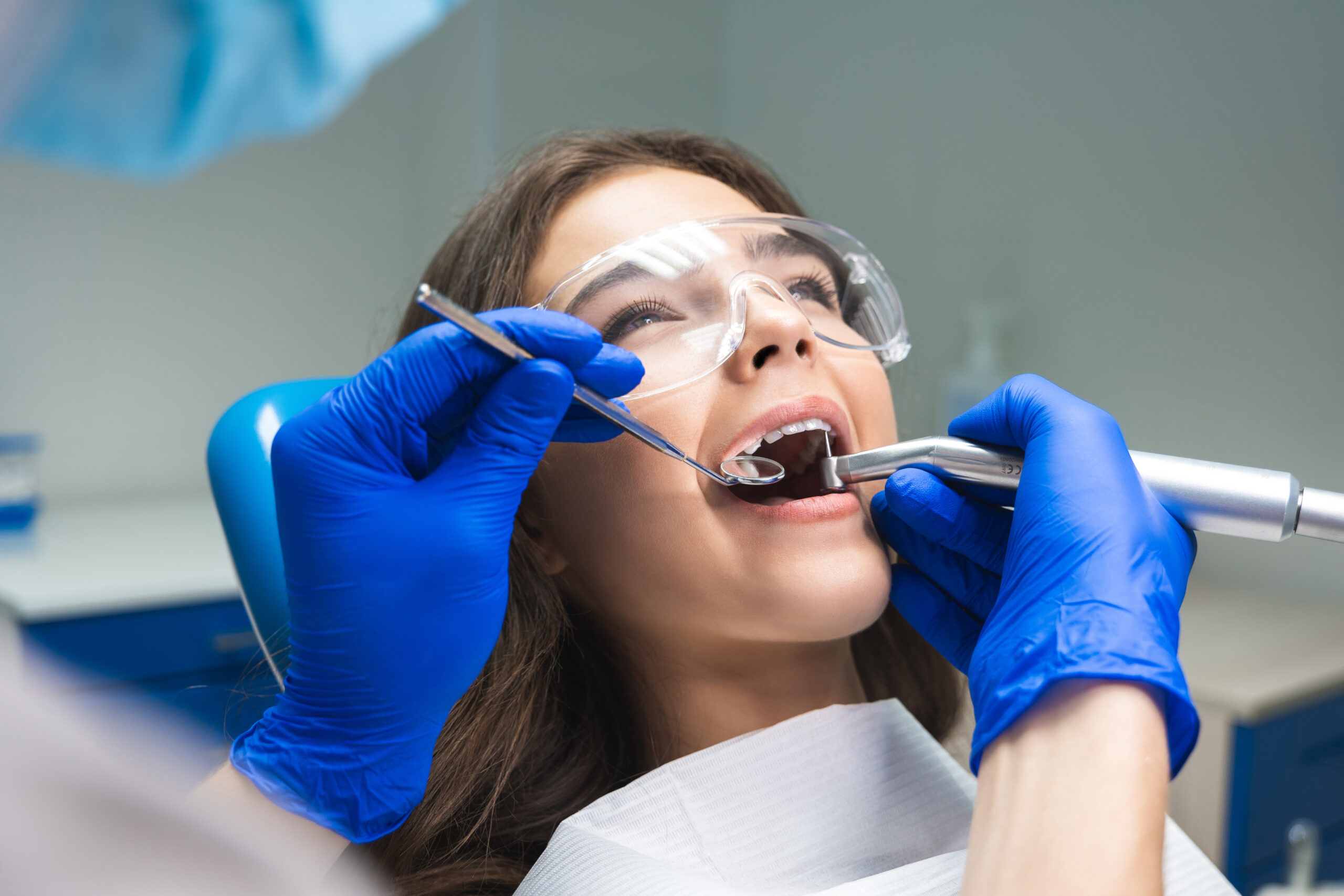 dentist in mask filling the patient's root canal while she is lying on dental chair wearing safety glasses under the medical lamp in dental office